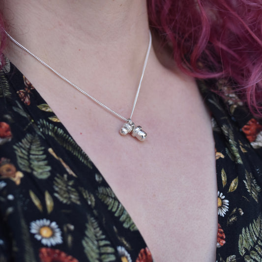 Girl with red hair and a dress with mushrooms and fliers is wearing a silver necklace with 2 different sized realistic acorns