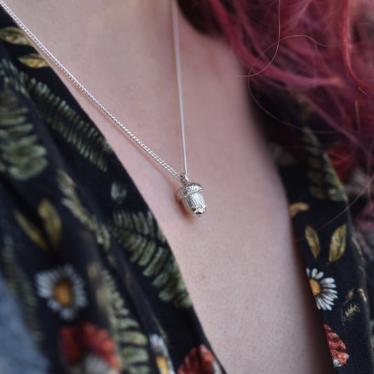 Girl with red hair and dress with mushrooms and flowers on is wearing a silver acorn pendant. 