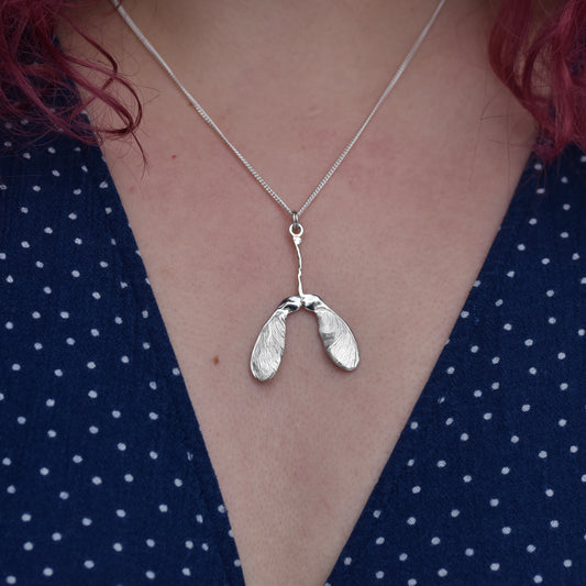 Girl wearing a blue dress is wearing a sycamore seed pendant. The pendant is highly detailed and sparkly with engraved details on a silver chain 