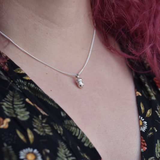 Girl with red hair wearing a dress with mushrooms and flowers on is wearing a silver pendant. The pendant is a small realistic acorn 