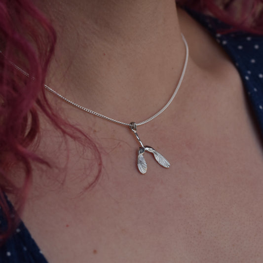 Girl wearing a blue dress, with red hair is wearing a sycamore seed pendant that is embellished with engraving and highly polished silver 