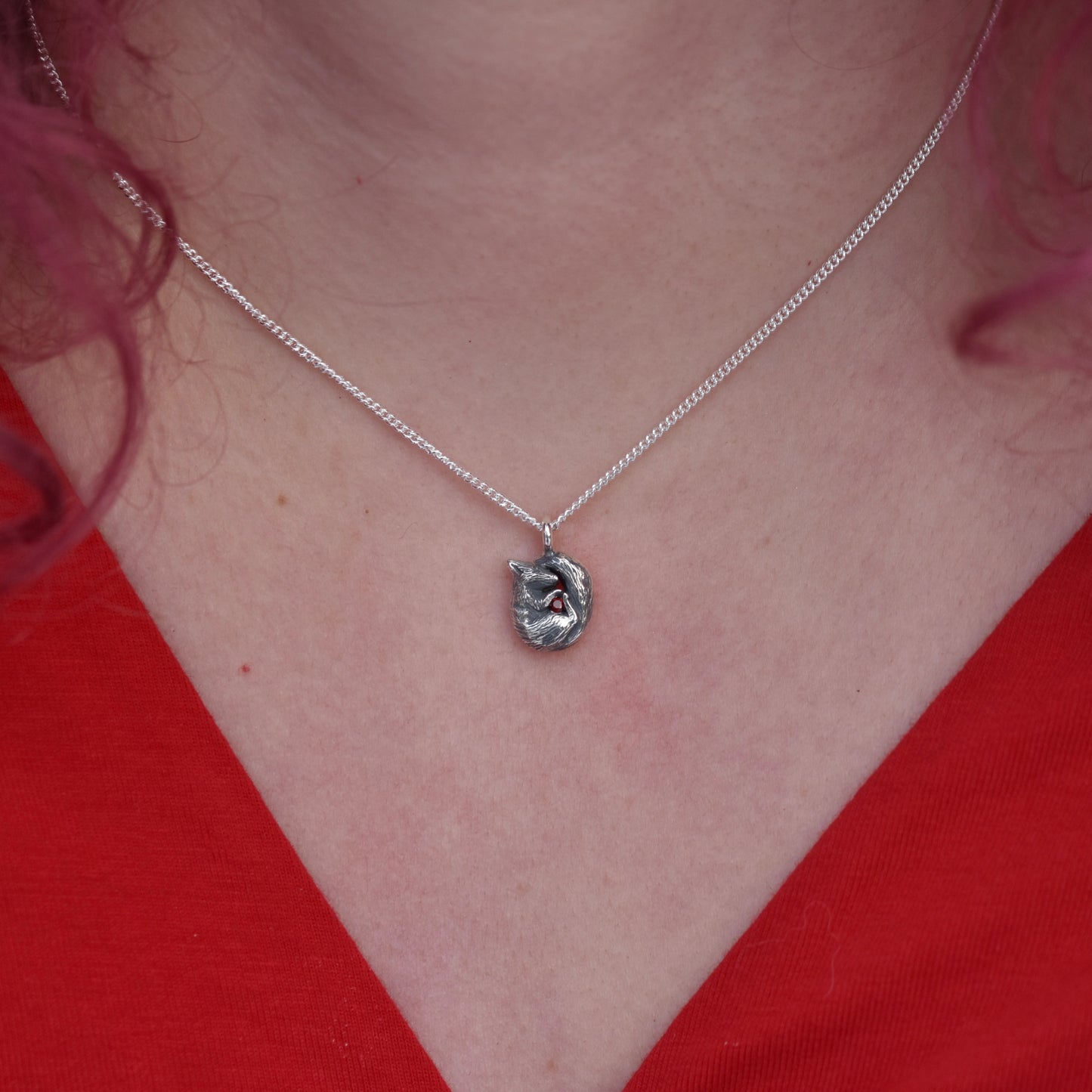 Girl with red hair and v neck ed top on is wearing a pendant made of silver. The pendant is a tiny curled up squirrel clutching a red gemstone which is a garnet 